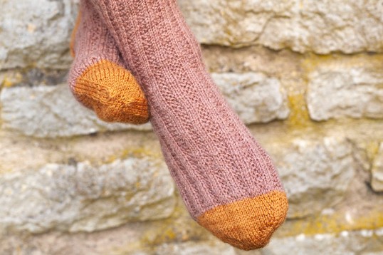 Färgfotografi av hemstickade raggsockor i rosa och gult mot en ljus stenmur.
