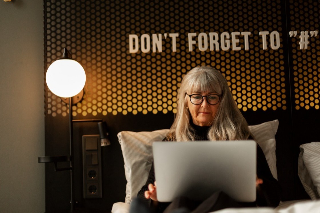 Kvinna med långt, grått hår och glasögon sitter i hotellsäng med laptop i knät.
