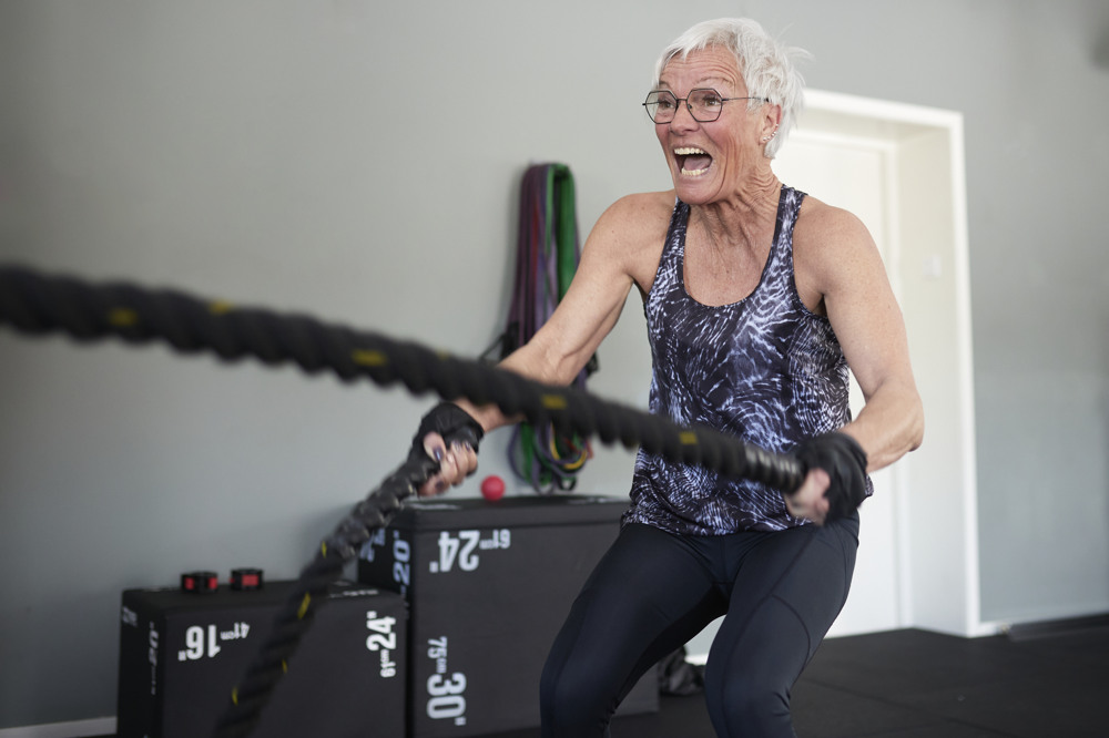 Vithårig kvinna med glasögon iklädd svarta träningskläder svingar rep på ett gym med stor inlevelse.