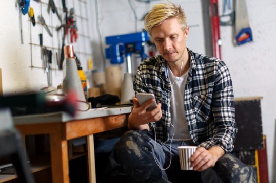 En företagare tar en paus i verkstaden och kollar mobilen. I andra handen, en kaffemugg.