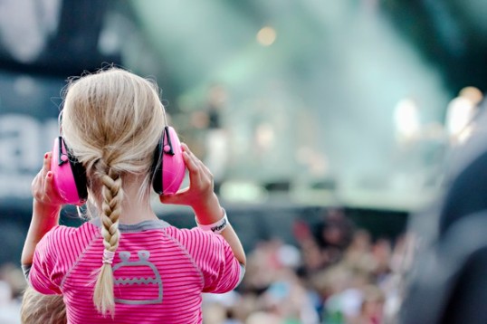 Färgfotografi av ett barn i konsertmiljö. Barnet är iklädd rosa tröja och har på sig knallrosa hörselskydd.