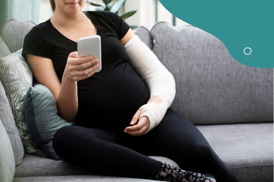 En gravid kvinna i svarta kläder sitter i en soffa. Vänster arm är gipsad, höger hand håller en telefon.